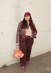 Brauner Lederblazer über einem beigefarbenen Pullover, braune Cordhosen, hält eine hellrosa Tasche, trägt eine kastanienbraune Mütze und burgunderrote Turnschuhe.