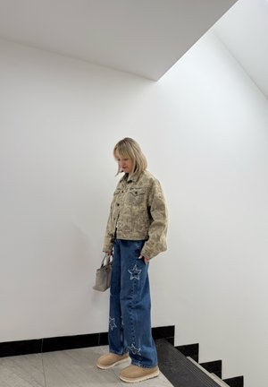 Chaqueta de mezclilla con estampado de camuflaje, jeans de pierna ancha azul con parches de estrellas, botas de plataforma beige y un pequeño bolso gris sobre un fondo blanco.