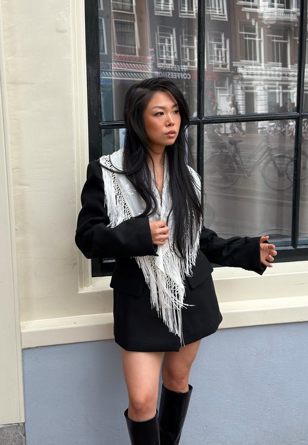 Femme aux longs cheveux noirs portant un manteau noir avec des détails en franges blanches et des bottes noires, debout devant une fenêtre dans une rue de la ville.