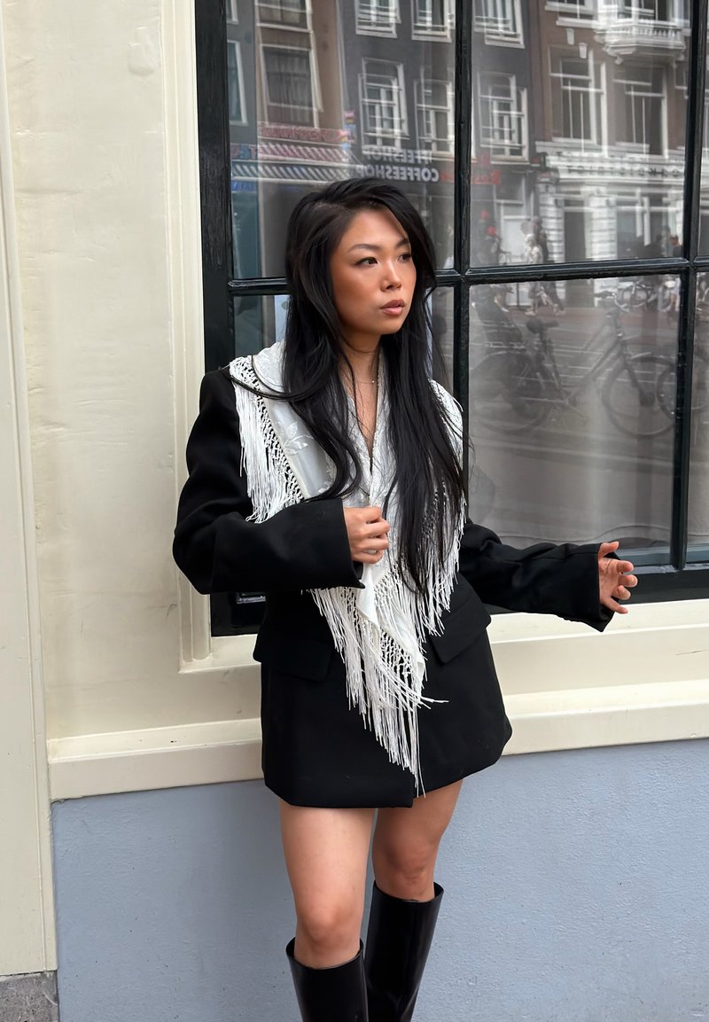Femme aux longs cheveux noirs portant un manteau noir avec des détails en franges blanches et des bottes noires, debout devant une fenêtre dans une rue de la ville.