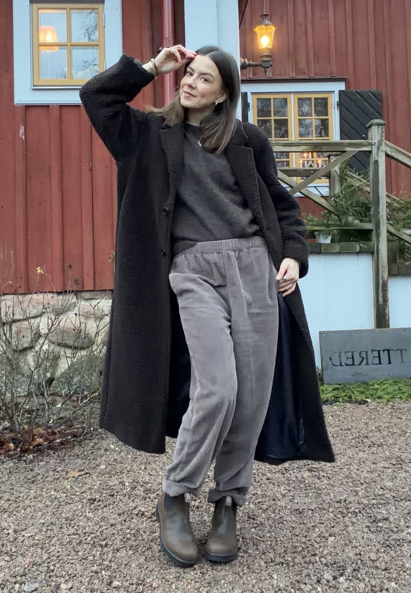 Sort, blød og lang frakke over en mørk sweater og lysegrå manchetbukser. Brune ankelstøvler. Udendørs miljø med rustik baggrund.