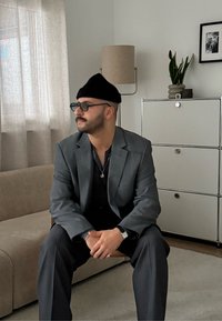 Costume gris avec une chemise noire, assis sur un canapé beige. Bonnet noir, lunettes rondes, montre argentée. Pièce neutre avec une lampe et une plante en pot.