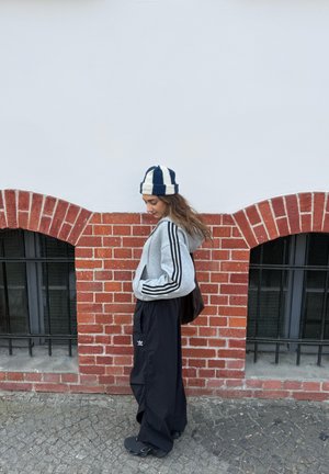 Joven mujer con pantalones anchos negros, sudadera gris con rayas y gorro de rayas, de pie contra un muro de ladrillos rojos con ventanas arqueadas.