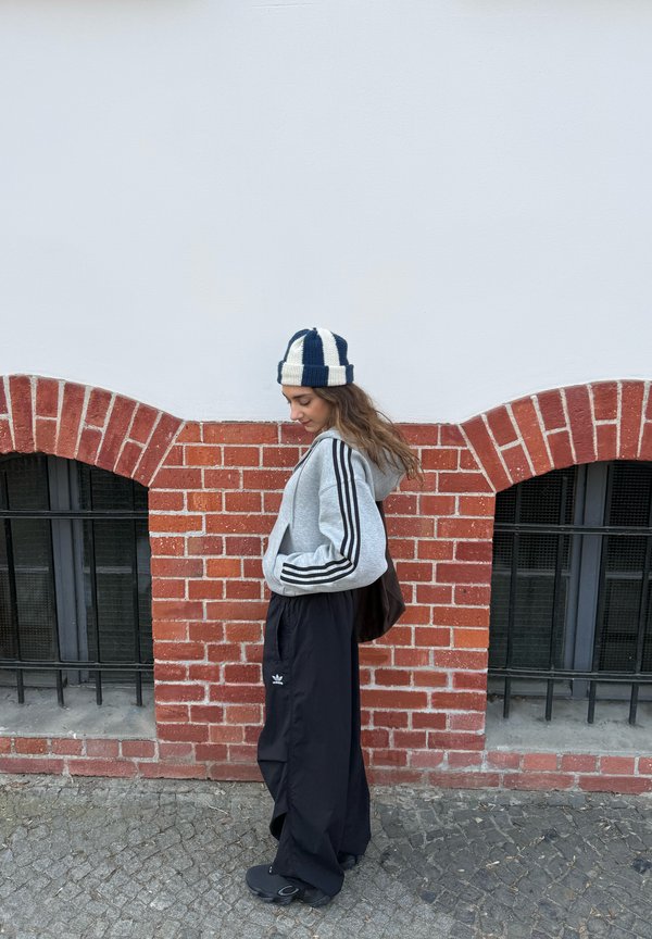 Jonge vrouw in zwarte wijde broek, grijze hoodie met strepen en gestreepte muts, staand tegen een rode bakstenen muur met ronde ramen.