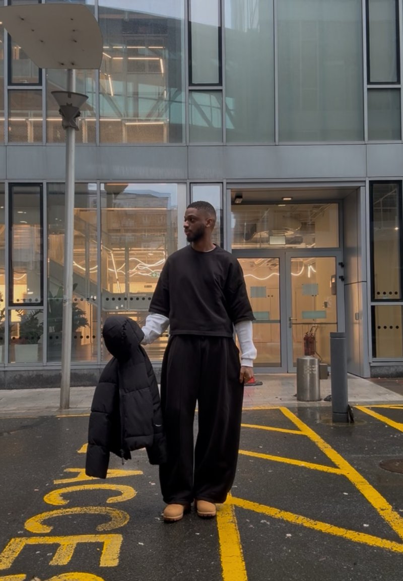 Homme vêtu d'une tenue noire tenant une doudoune noire, debout sur un bitume mouillé avec des lignes de stationnement pour personnes à mobilité réduite jaunes près d'un bâtiment moderne en verre.