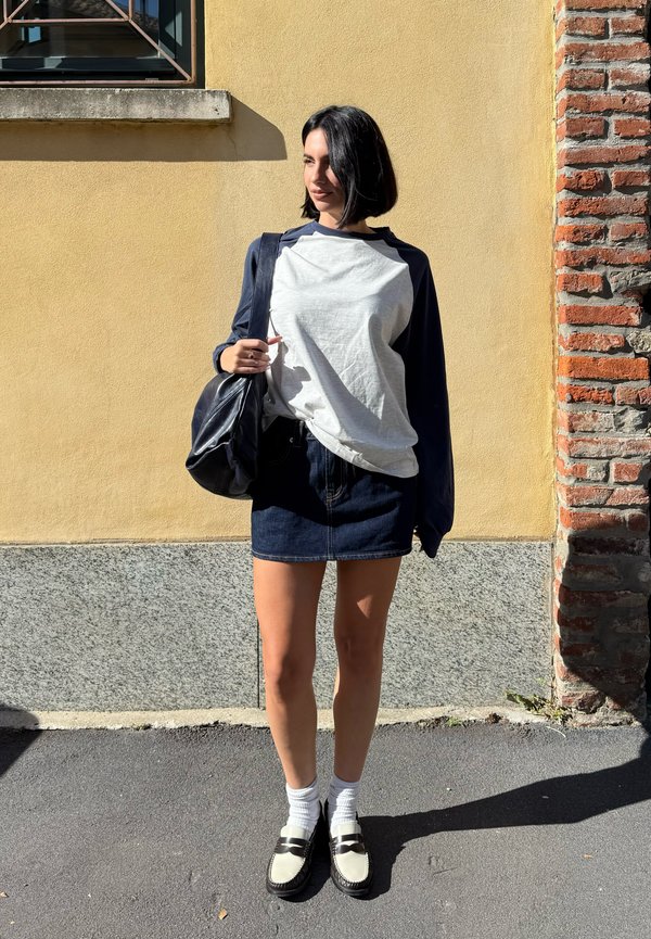 Une femme se tient au soleil devant un mur jaune. Elle porte un haut blanc et bleu marine, une jupe en jean, des chaussettes et des mocassins, ainsi qu’un sac noir à bandoulière.