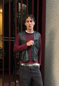 Gilet en cuir sur une chemise à manches longues rouge et noire à rayures, ceinture à étoile argentée, pantalon sombre ; debout contre un mur texturé avec des barres verticales.