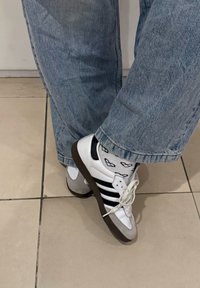 Osoba nosi svijetloplave traperice, bijele čarape s uzorkom crnih naočala i bijele Adidas tenisice s crnim prugama, stojeći na pločicama.