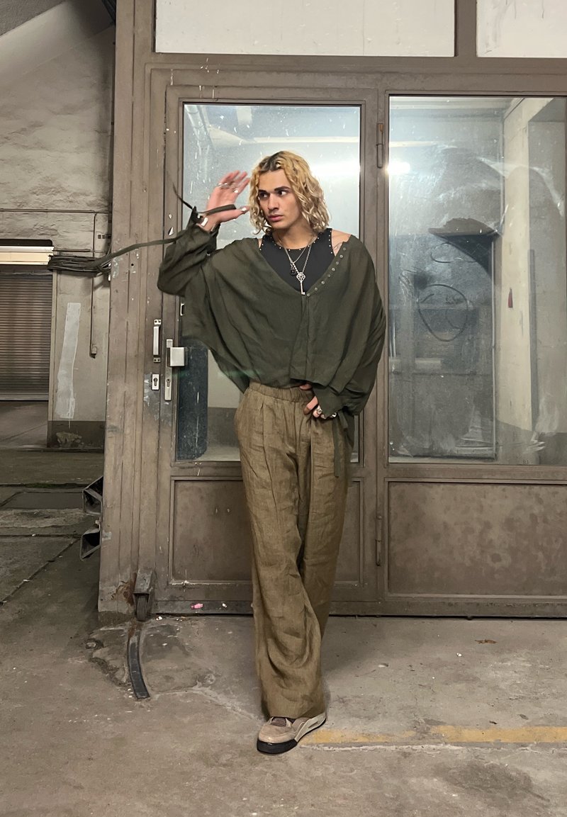 Overdeed olijfgroene shirt met knoopdetails, zwarte gelaagde top, losse beige linnen broek en grijze sneakers. Industriële achtergrond.