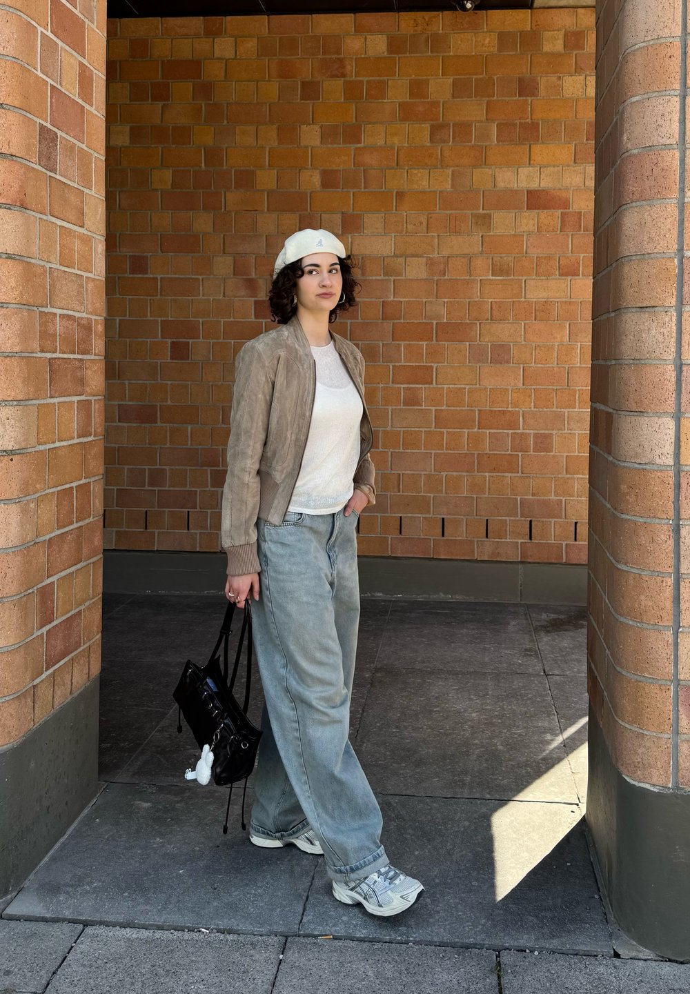 Jaqueta bomber em camurça em taupe, blusa de malha branca, jeans leves de cor azul claro, tênis cinzas, bolsa preta com um charme, de pé contra uma parede de tijolos.