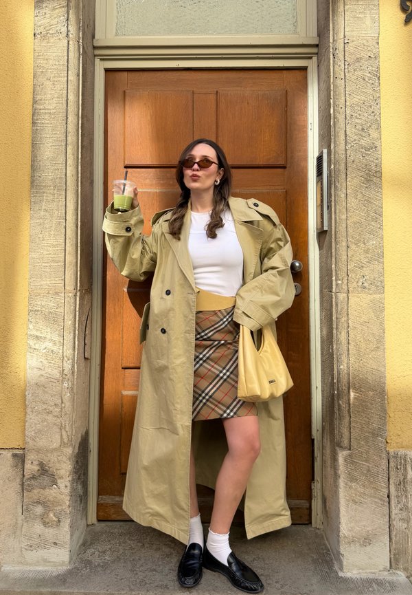Femme portant un trench-coat beige, une jupe à carreaux, un haut blanc, des lunettes de soleil, tenant un sac jaune et une boisson, debout devant une porte en bois.