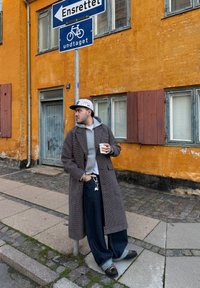 Abrigo de cuadros, sudadera gris con capucha, camiseta blanca, jeans anchos azules, zapatos oscuros, sosteniendo una taza. Pared naranja con señal de bicicleta y persianas de fondo.