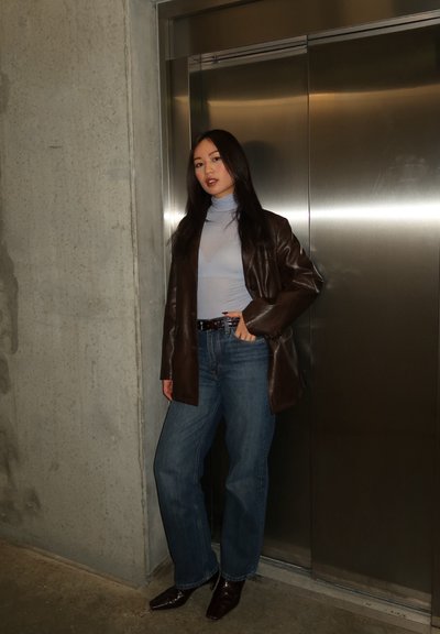 Mujer con cabello largo y oscuro, vestida con chaqueta de cuero marrón, suéter de cuello alto azul claro, jeans azules y zapatos oscuros, de pie junto a un ascensor de acero inoxidable.