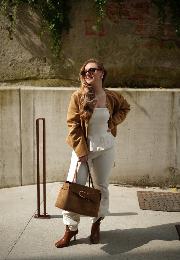 Kvinde iført brun jakke, hvid top og bukser, brune ankelstøvler, der holder en stor brun håndtaske, står udendørs ved en betonvæg.