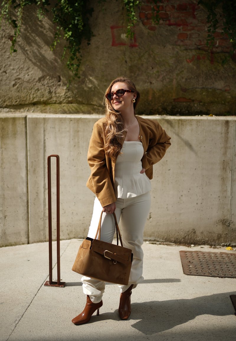 Femme portant une veste marron, un haut blanc et un pantalon, des bottines marron, tenant un grand sac à main marron, debout à l'extérieur contre un mur en béton.