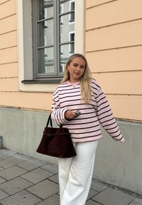 Langærmet, lyserød stribet sweatshirt lagdelt over hvide bukser med vidde i benene. Brun ruskindshåndtaske med struktureret form. Lys baggrund med et vindue.
