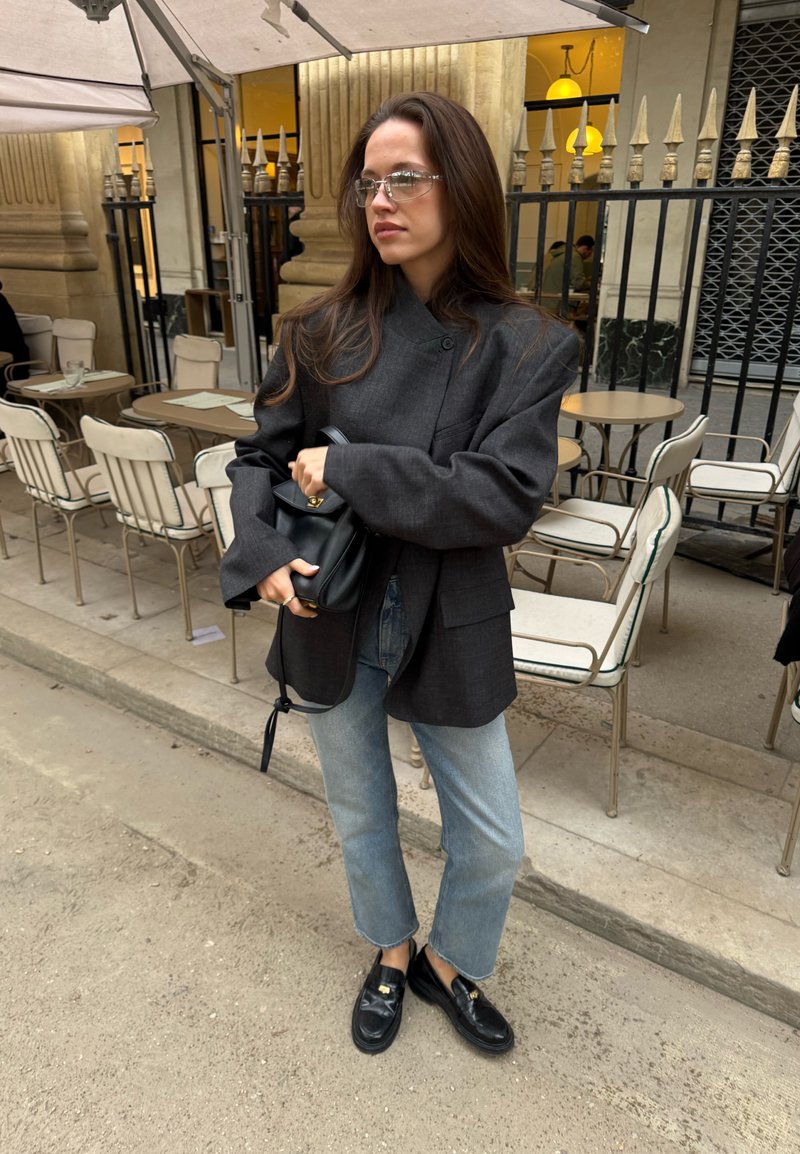 Black oversized blazer, light blue straight-leg jeans, shiny black loafers, clear frames on glasses, holding a small black bag.