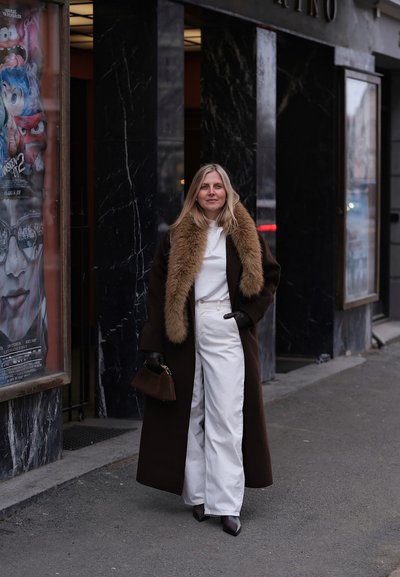 Mujer con abrigo largo y oscuro con cuello de piel, pantalones blancos, guantes negros y botas, de pie en la acera de la ciudad cerca de carteles de películas.