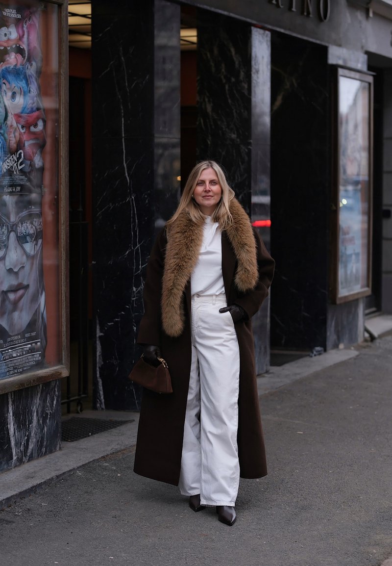 Γυναίκα με μακρύ σκούρο παλτό με γούνινο γιακά, λευκό παντελόνι, μαύρα γάντια και μπότες, στέκεται σε πεζοδρόμιο της πόλης κοντά σε αφίσες ταινιών.