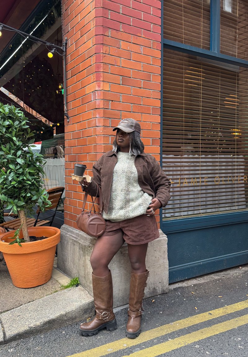 Person wearing brown jacket, shorts, boots, and cap, holding coffee next to brick wall outside café with large plant in pot nearby.