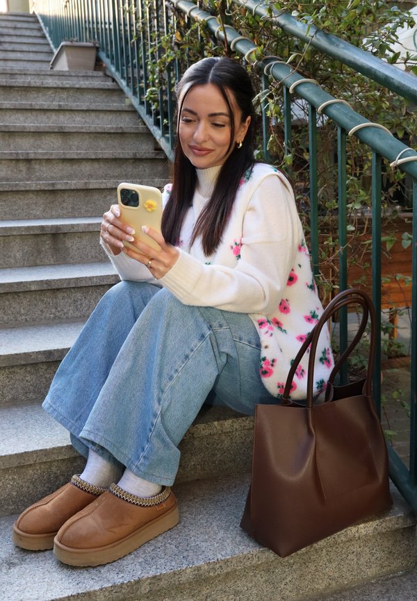 Femeie așezată pe scări în aer liber, purtând un vestă tricotată cu flori și blugi albaștri, uitându-se la smartphone cu o geantă maro din piele lângă ea.