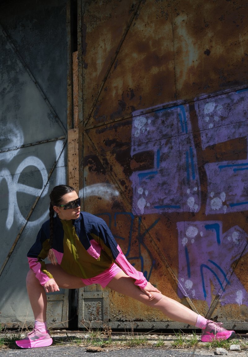 Femme en tenue de sport colorée et lunettes de soleil étirant la jambe en plein air devant une porte métallique rouillée avec un graffiti violet.