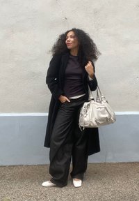 Mulher com cabelo encaracolado vestindo um casaco preto, calças largas escuras e sapatos brancos, segurando uma grande mala branca, de pé contra uma parede texturizada.