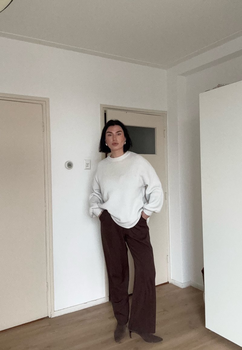 Femme aux cheveux noirs debout à l'intérieur, portant un pull blanc, un pantalon large marron et des bottes à talons marron, les mains dans les poches.