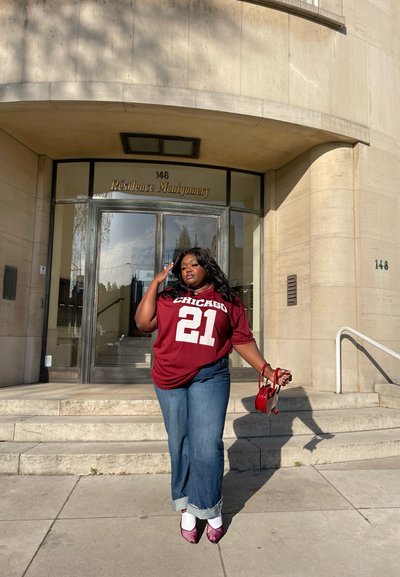 Jersey de gran tamaño en color marrón con "CHICAGO" y "21", combinado con pantalones de mezclilla anchos en azul claro. Sosteniendo un pequeño bolso rojo, de pie en escalones de concreto.