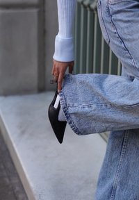 Sort højhælet sko med spids tå og åbne sider, båret med blå, slidte vidde-ben jeans og hvide sokker. Fokus på tekstildetaljer og pasform.