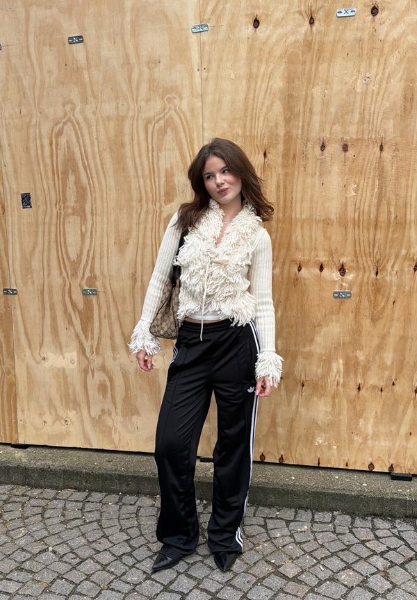 Jonge vrouw staat op een geplaveide straat en draagt een witte, gestructureerde trui, zwarte sportbroek met witte strepen en zwarte puntige schoenen.