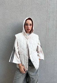 White quilted vest with a hood, layered over a ribbed beige top with black stripes. Olive green relaxed-fit trousers.