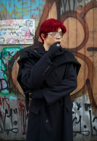 Schwarzer Mantel mit großen Revers und Knöpfen, kombiniert mit einem schwarzen Schal. Rote Haare, silberne Sonnenbrille und schwarze Handschuhe mit detaillierten Akzenten. Hintergrund mit Graffiti.