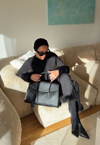 Sac à main en cuir noir avec une poignée supérieure et un détail à franges, posé sur les genoux. Le modèle porte une tenue noire avec des poignets en fourrure et des chaussures noires élégantes.