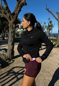 Haut de sport noir à manches longues avec une demi-fermeture éclair, associé à un short bordeaux doté d'une poche latérale contenant un objet rose. En arrière-plan, des arbres.