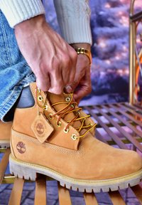 Bottes en daim marron clair avec des lacets jaunes et rouges, des œillets dorés et une semelle en bois. Présente une étiquette indiquant "Étanche garanti" avec un logo.