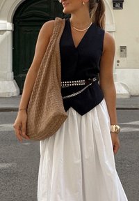 Schwarzes ärmelloses Blusenshirt, weiße fließende Rock, gewebte beige Tasche, geschichtete Halsketten, silberner Gürtel mit Nieten und eine Uhr am Handgelenk.