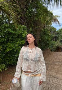 Blouse blanche transparente avec broderie florale, manches longues et lien au col, associée à un pantalon ample crème, tenant une tasse.