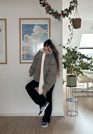 Chaqueta acolchada verde oliva con cuello de forro polar blanco, pantalones negros y zapatillas azules. Ambiente interior con una planta y una obra de arte enmarcada.