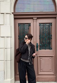 Musta tikattu takki, harmaa pusero, mustat housut ja aurinkolasit. Seisoo ruskean oven vieressä, jossa on turkoosit koristeelliset yksityiskohdat ja lasipaneelit.