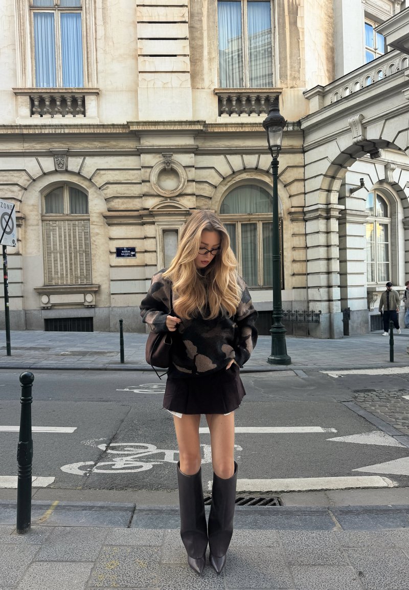 Ung kvinne med langt blondt hår, iført briller, en mønstret genser, svart skjørt og høye svarte støvler, stående på en byfortau.