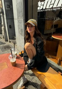 Blazer oversized preto, boné estampado com um padrão de chevron amarelo e laranja, meias brancas, ténis cinzentos. Duas bebidas geladas numa mesa de madeira.