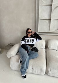 Μαύρη και άσπρη κομμένη φούτερ FUBU, ανοιχτό μπλε φαρδιά τζιν, μαύρα αθλητικά παπούτσια με λευκές λεπτομέρειες, καθισμένος σε έναν ανοιχτό γκρι αφράτο καναπέ.