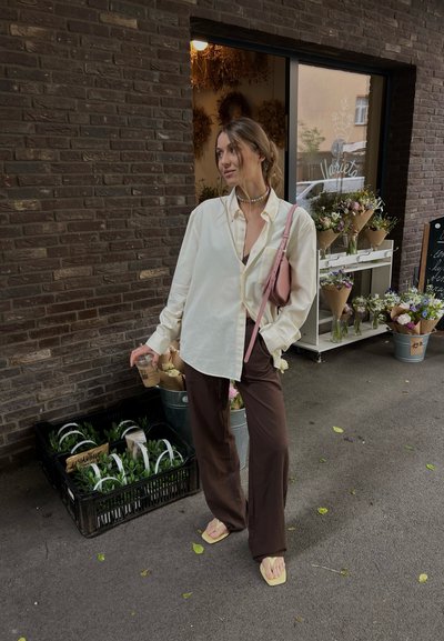 Camisa blanca de gran tamaño con botones, pantalones anchos marrones y chanclas amarillas. Mujer sostiene una bebida, de pie junto a arreglos florales.