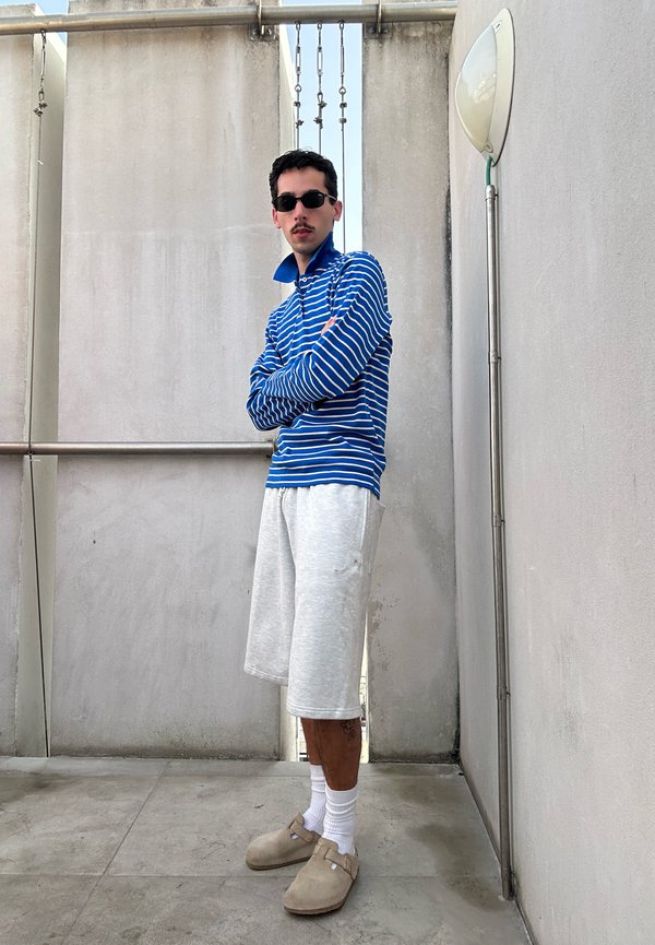 Άντρας που φορά γυαλιά ηλίου, μπλε και άσπρη ριγωτή μπλούζα με μακριά μανίκια, γκρι σορτς, λευκές κάλτσες και μπεζ παπούτσια slip-on, στέκεται με σταυρωμένα τα χέρια.