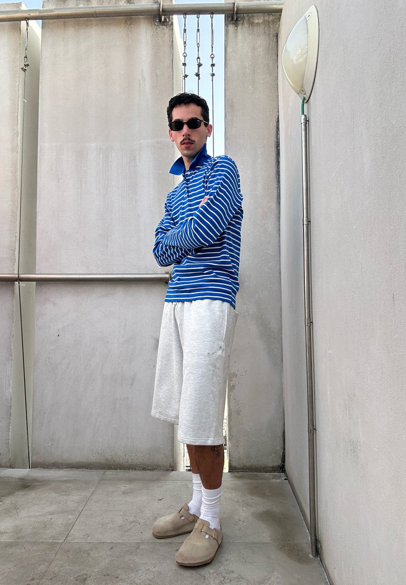 Άντρας που φορά γυαλιά ηλίου, μπλε και άσπρη ριγωτή μπλούζα με μακριά μανίκια, γκρι σορτς, λευκές κάλτσες και μπεζ παπούτσια slip-on, στέκεται με σταυρωμένα τα χέρια.