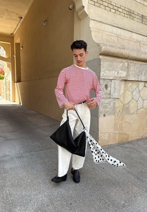 Joven con camisa de rayas rojas y blancas y pantalones blancos, sosteniendo una gran bolsa negra con un pañuelo de lunares blanco y negro atado a ella.