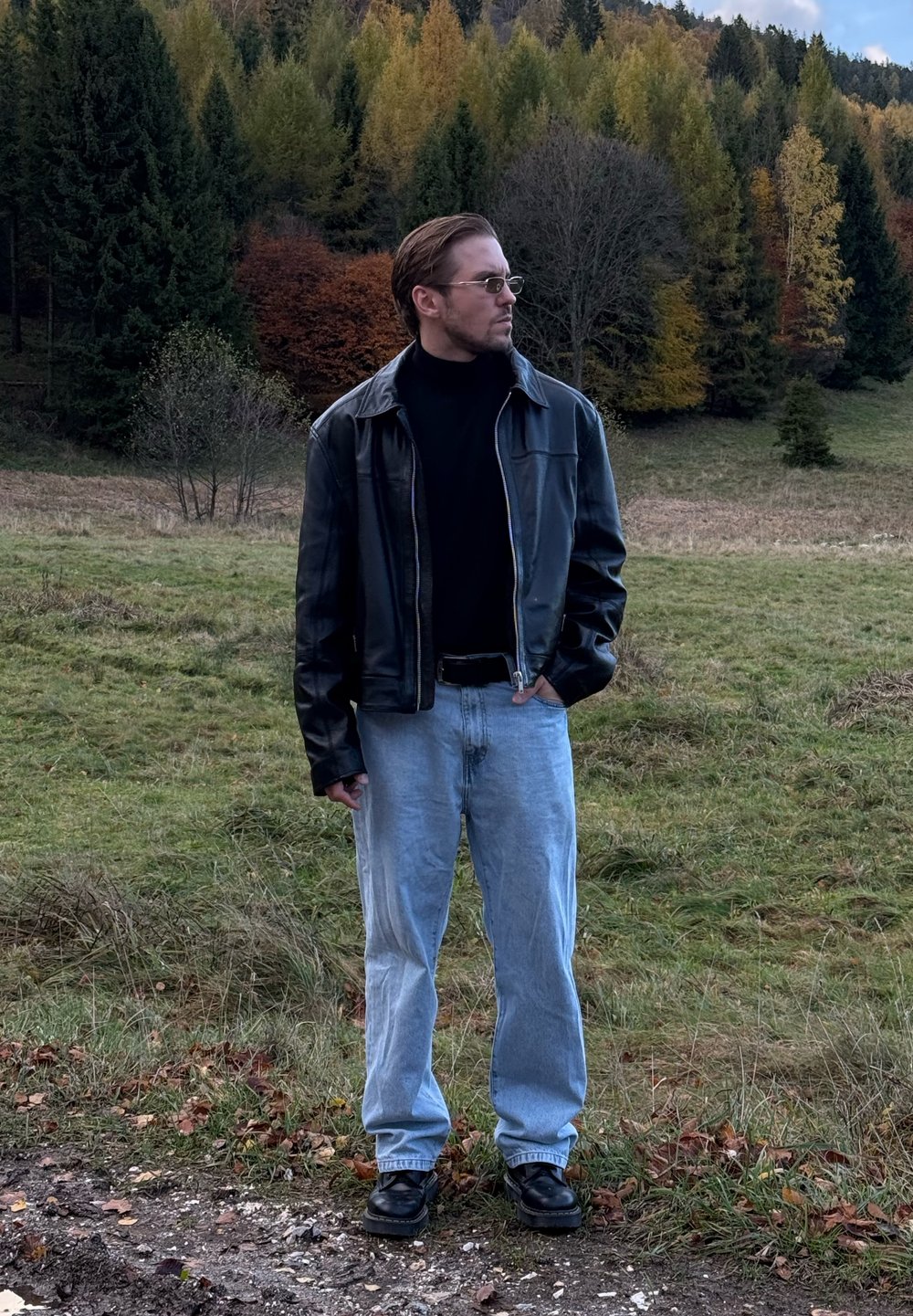 Zwarte leren jas over een zwarte coltrui, lichtblauwe spijkerbroek en zwarte schoenen; staand in een grasachtig gebied met bomen op de achtergrond.