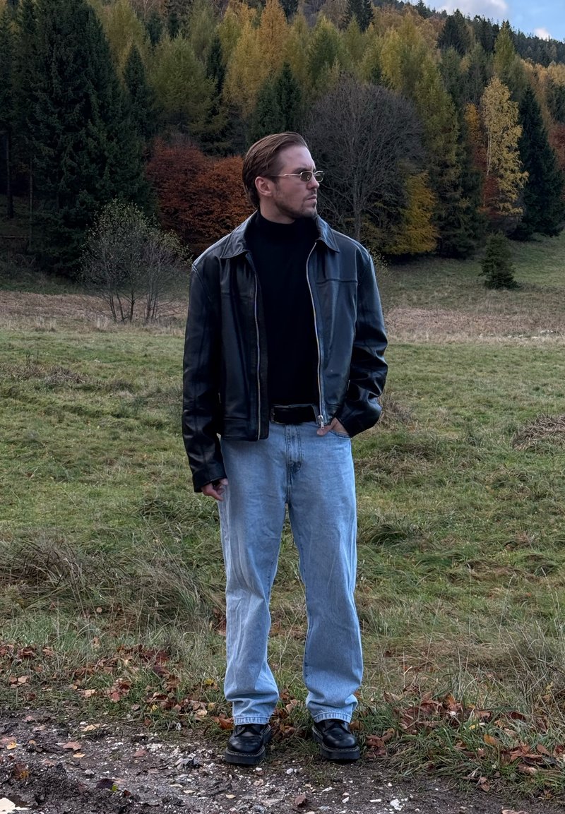 Giacca di pelle nera sopra un dolcevita nero, jeans azzurri e scarpe nere; in piedi in un'area erbosa con alberi sullo sfondo.