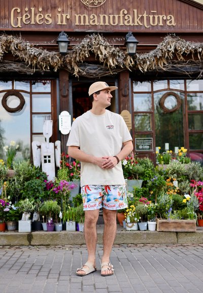 Camiseta de manga corta beige, pantalones cortos tropicales coloridos y sandalias ligeras. El fondo presenta varias flores en macetas y una tienda de madera.